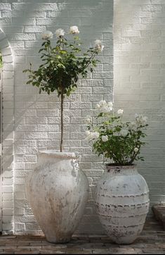 two white vases with flowers in them sitting on a brick floor next to a wall