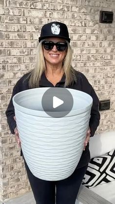 a woman holding a large white bucket in front of a brick wall and wearing sunglasses