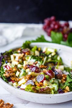 a salad in a white bowl with grapes and walnuts on the table next to it