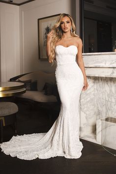 a woman in a white dress standing next to a fireplace with her hand on her hip