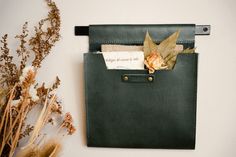 a green leather case with some flowers and papers in it on a white wall next to dried plants