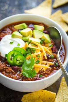 a white bowl filled with chili, cheese and avocado next to tortilla chips