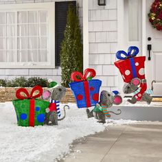 two metal mice carrying presents in front of a house with snow on the ground and wreaths around them