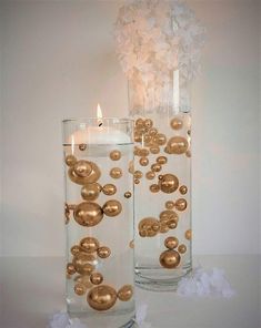 two clear vases with gold balls and a lit candle are on a white table