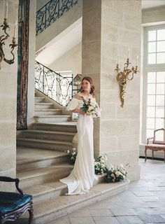 a woman in a wedding dress standing on the stairs with her bouquet and looking at the camera