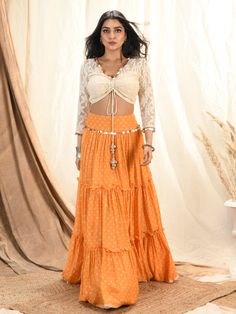 This is a three-piece mango bandhni lehenga set from the Aaryaa by KK collection. This beautiful silk mul tiered lehenga is paired with a floral lace blouse. The outfit is completed with a dupatta with coins tassel edging and it’s crafted in crush tissue material. The mango color with bandhni pattern, shells, and coin tassels enhance this lehenga set. The front tie-up style shimmer blouse has a V neckline with coin tassel embellishments and a back opening. Designer Ruffled Lehenga For Navratri, Designer Tiered Lehenga With Dupatta, Designer Lehenga With Ruffles For Navratri, Eid Floor-length Ruffled Lehenga, Bollywood Style Ruffled Lehenga For Navratri, Designer Festive Tiered Skirt Lehenga, Anarkali Choli With Dupatta And Tiered Skirt, Designer Festive Lehenga With Tiered Skirt, Bollywood Style Choli With Dupatta And Tiered Skirt