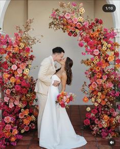 a bride and groom kissing in front of an archway with flowers on the wall behind them