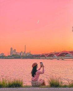 a woman sitting on the edge of a river taking a photo with her cell phone