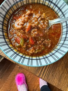 a bowl of chili and meat soup with someone's feet on the table next to it