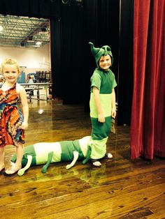 two children in costumes standing next to a fake lizard on the floor with red curtains behind them