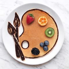 a pancake decorated with fruit and chocolate on a white plate next to utensils