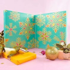 an open christmas card with presents and decorations on the table next to it, in front of a pink background