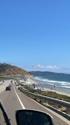 cars driving down the road next to the beach and ocean on a sunny day with blue skies