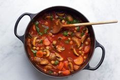 a pot filled with stew and vegetables on top of a white counter next to a wooden spoon