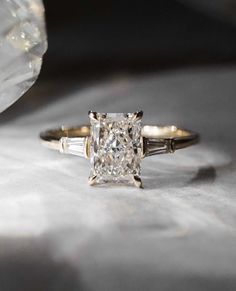 an engagement ring with a princess cut diamond and baguettes on the side, sitting on a white surface