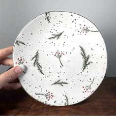 a hand holding a white plate with red berries and green leaves on it, sitting on a wooden table