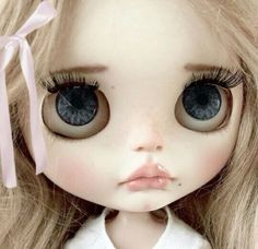 a doll with long blonde hair and big blue eyes wearing a white shirt, pink ribbon around her neck