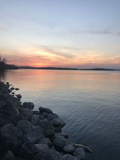 the sun is setting over the water and rocks are in the foreground, as seen from the shore