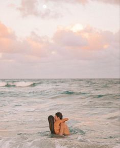 a couple kissing in the ocean with waves crashing on them and pink clouds behind them