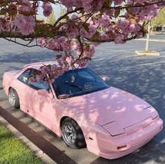 a pink sports car parked under a tree with flowers on it's branches in front of a parking lot