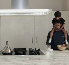 a man holding a young child while standing in front of a stove top with a bowl on it