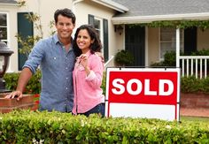 a man and woman standing next to a sold sign in front of a house with bushes