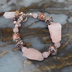 "The bracelet has 2 large rough quartz stones (ab. 2.5 x 3 cm /1\" x 1.2\"), 8 mm round pink quartz beads, 2 10  mm Leopard Skin jasper beads, metal beads and caps. The closure is with a hook, alongside which hangs a cluster consisting of a pink quartz obelisk (brass cap, 36x11x11 mm /1.42\" x 0.4\" x 0.4\"), 2 quartz beads, and 4 freshwater pearls (3 light pink, 1 black). The carrying cord is tigertail stainless steel. The bracelet is 21 cm /8.3\" long and weighs 70 gr. / 2.47 oz. Rose quartz is known as a healing crystal and the stone of unconditional love. It restores trust and harmony in relationships, purifies and opens the heart at all levels to promote love, self-love, friendship, deep inner healing and feelings of peace. Rose Quartz dispels negativity and encourages self-forgivenes Handmade Statement Crystal Bracelet, Luxury Pink Rose Quartz Jewelry, Luxury Rose Quartz Jewelry With Natural Stones, Luxury Rose Quartz Natural Stones Jewelry, Raw Stone Charm Bracelet, Luxury Handmade Crystal Bracelet, Women's Rose Quartz Bracelets, Rough Stone Bracelet, Spiritual Rose Quartz Jewelry With Round Beads