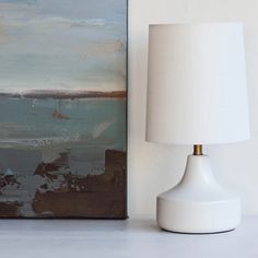 a white lamp sitting next to a painting on a wall with a light shade over it