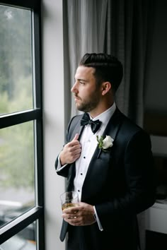 a man in a tuxedo standing by a window with a drink in his hand