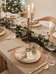 the table is set for christmas dinner with candles and greenery on it, along with place settings