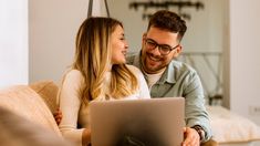 a man and woman sitting on a couch looking at a laptop computer screen while smiling