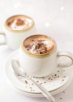 two white cups filled with hot chocolate on top of saucers next to spoons