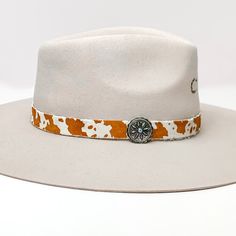 Cow Print Hat Band with Silver Tone Charm in Brown, and Ivory. Pictured on a white background with the band around a light tan/ brown hat. Adjustable Felt Hat For Kentucky Derby With Short Brim, Kentucky Derby Adjustable Short Brim Felt Hat, Adjustable Fedora For Country Events, Adjustable Wide Brim Felt Hat For Country Events, White Adjustable Fedora With Flat Crown, Adjustable White Fedora With Flat Crown, Adjustable Wide Brim Country Style Top Hat, Adjustable Wide Brim Top Hat For Ranch, Adjustable Wide Brim Top Hat For Country Events