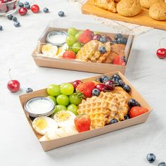 two boxes filled with different types of food on top of a white table covered in blueberries, strawberries and other fruit