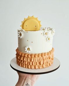 a hand holding a cake decorated with flowers and sun on top, in front of a white background