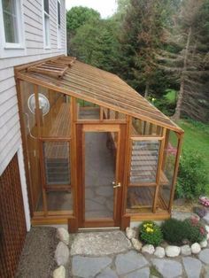 a small wooden greenhouse with glass doors on the front and side of it, next to a house