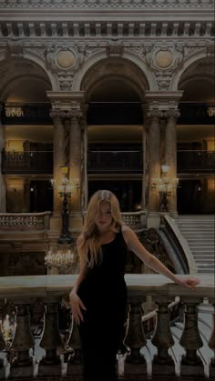 a woman in a black dress is standing on a balcony with chandeliers and stairs