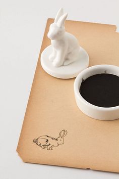 a white rabbit figurine sitting on top of a piece of paper next to a cup of coffee