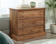 a wooden dresser sitting next to a window with books on top of it and a potted plant in the corner