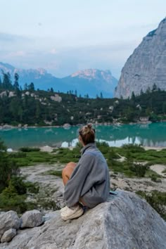 a person sitting on top of a large rock next to a body of water with mountains in the background