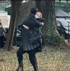 two people hugging each other in front of trees and parked cars on the side of the road