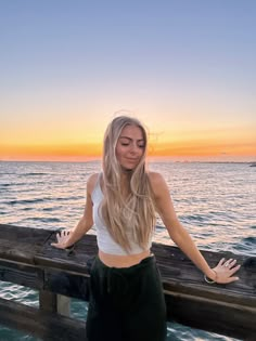 a beautiful young woman standing on top of a wooden pier next to the ocean at sunset