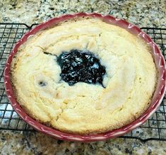 a baked cake with blueberry topping in a red bowl on a wire rack next to a marble counter top