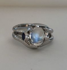 The Crystal Ball Ring is a very unique design, crafted by Heirloom Pavé in 18K white gold, featuring a Cabochon Moonstone. And two perfectly blue round brilliant Sapphires. The Moonstone is a oval shape Cabochon with a beautiful iridescent that so mystical and other worldly looking. The two, bezel-set Sapphires are a total carat weight of .35ct are a beautiful accent to the center. In the high polished white gold it is a true statement piece. Size 7 (Sizable upon request) Every Antique piece of Silver Moonstone Ring With Stone Setting, Unique Silver Moonstone Ring With Stone Setting, Silver Fusion Moonstone Ring, Silver Fusion Style Moonstone Ring, Silver Fusion Style Moonstone Gemstone Ring, Silver Moonstone Gemstone Ring, Sterling Silver Cabochon Moonstone Ring For Wedding, Wedding Sterling Silver Cabochon Moonstone Ring, Unique Sterling Silver Moonstone Cabochon Ring