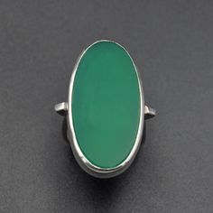 This beautiful modernist ring features an elongated oval shaped green chalcedony, bezel set in silver. The hue of the gem is lovely: a bright and earthy green. The shoulders of the ring are slightly flared, then taper and narrow through the square shaped shank, flaring again slightly at the flattened base. The band of the ring is squared but fits comfortably on the finger. The back of the gallery has a textured motif. Chalcedony is a crypto-crystalline variety of quartz.   More details: Central chalcedony cabochon in bezel measures 31.3mm tall and 17.0mm wide. Setting rises 5.5mm off the finger. At back of ring, ring shank measures 3.9mm. Ring size 6 / UK M. Due to the design of this ring with its square shank, resizing is not possible.  Excellent condition Sterling silver (stamped.) 11.2 Modern Green Ring With Large Stone, Modernist Oval Cabochon Rings, Modernist Oval Gemstone Rings, Modern Green Oval Cabochon Jewelry, Oval Green Onyx Gemstone Ring, Oval Gemstone Rings In Modernist Style, Modern Green Cabochon Rings, Oval Green Onyx Rings, Modern Oval Green Jewelry