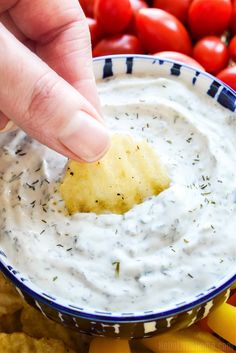 a hand dipping a tortilla chip into a bowl of ranch dip with tomatoes in the background