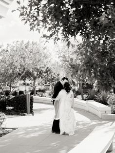 a bride and groom standing on the sidewalk in front of some trees with their arms around each other