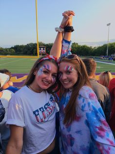 two girls with face paint on their faces at a football game, one holding up the other's hand