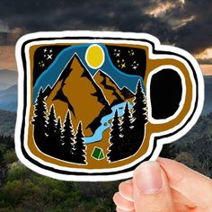 a hand holding up a sticker with mountains and trees in the background at sunset