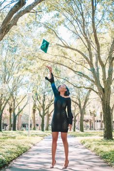 a woman in a black dress is flying a green kite through the air with trees behind her